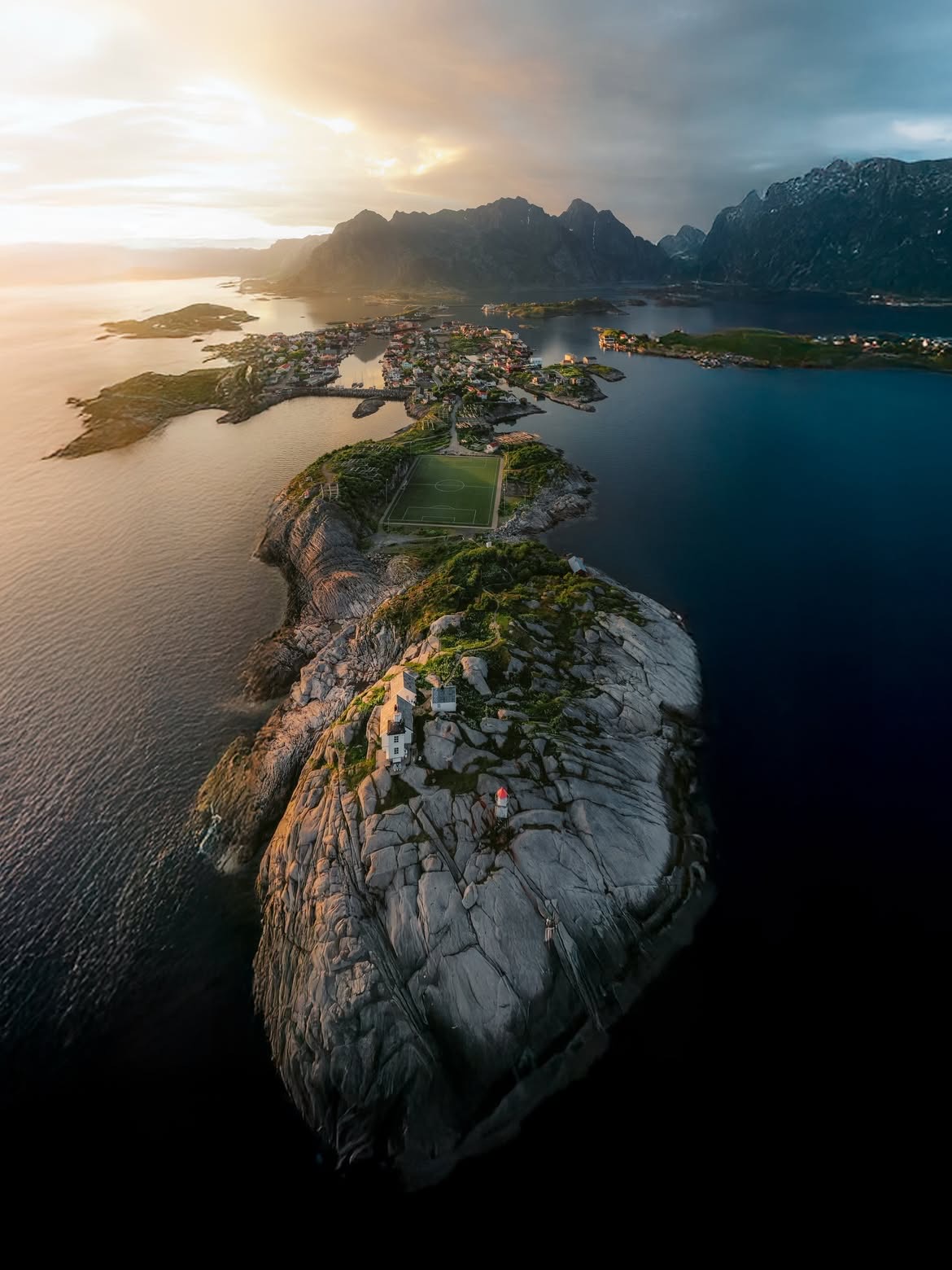 Aerial view of Henningsvaer at sunset. A green football pitch sits on a bare rock island ringed by sea, with the fishing village and the Lofoten mountains behind it